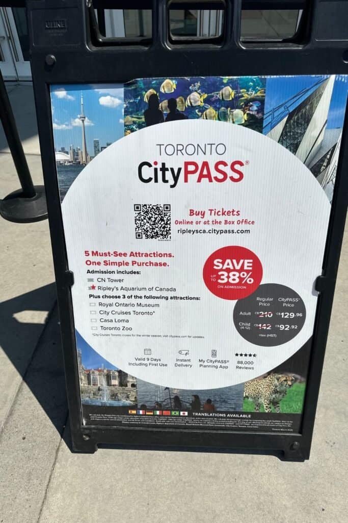 A promotional sign for the Toronto CityPASS displayed on a sidewalk. The sign highlights that the CityPASS includes admission to CN Tower and Ripley’s Aquarium of Canada, plus a choice of 3 additional attractions from the Royal Ontario Museum, City Cruises Toronto, Casa Loma, and the Toronto Zoo. The sign states that CityPASS saves up to 38% on admission costs, with adult tickets priced at $129.96 CAD and child tickets at $92.92 CAD. The pass is valid for 9 days, offers instant delivery, and includes a planning app. Images of the CN Tower, aquarium, museum, and other attractions are featured. A QR code and website URL (ripleysca.citypass.com) are also visible.